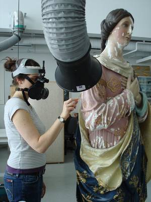 Photographie d'une restauratrice procédant au nettoyage d'une madone sous le système d'extraction d'air et muni d'un masque à filtre HEPA. 